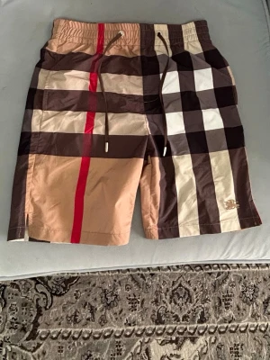 Rutig shorts med snörning  - Snygga shorts med klassiskt rutigt mönster i beige, brunt, svart, vitt och en röd rand. Shortsen har elastisk midja med snörning och en bakficka med knapp. Perfekta för sommaren och ger en trendig look och kan användas som bad shorts.
