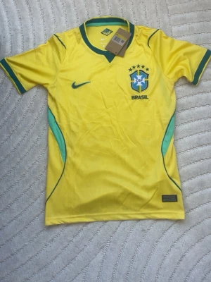 Gul Brasil Nike fotbollströja - Säljer en gul Brasilien fotbollströja från Nike med gröna detaljer och CBF-emblem på bröstet. Helt ny och har taggen kvar. Passar S men också xs