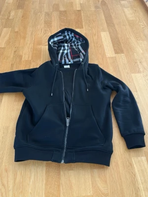 Svart hoodie med dragkedja och mönstrad huva helt ny  - Snygg svart hoodie med dragkedja och två stora fickor framtill. Huvan har ett rutigt innerfoder i svart, vitt och rött, samt dragsko. Perfekt för en avslappnad och cool stil.