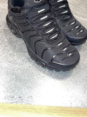Nike Air Max TN skor - Nike Air Max Plus sneakers i helsvart med mesh- och syntetmaterial. Karaktäristiska vågformade detaljer och synlig Air-dämpning i sulan. Tjock yttersula med bra grepp och TN-logga undertill. Perfekta för dig som gillar sportig och clean stil.