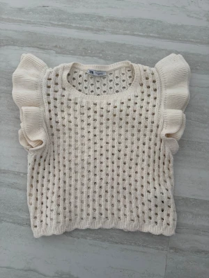 Vit stickad topp med volangärm Zara - Söt stickad topp från Zara i offwhite med hålmönster och volangärmar. Rund halsringning och kort ärm ger en trendig look. Perfekt att styla till jeans eller kjol för en fräsch outfit. Materialet är mjukt och luftigt, vilket gör den skön att bära.