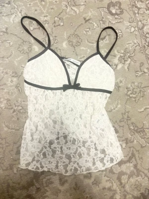 Vit spets med svarta detaljer Bershka - Säljer en vit spetsbody från Bershka med tunna axelband och svarta kanter. Bodyn har en liten rosett framtill och är helt i blommig spets som ger en romantisk vibe. Perfekt för dig som gillar feminina detaljer och transparent look.