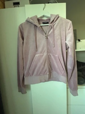 Ljusrosa velour hoodie från Juicy Couture - Säljer en ljusrosa hoodie i mjuk velour från Juicy Couture. Hoodien har dragkedja framtill, två fickor och justerbar huva med snören. Ikonisk JC-logga broderad på bröstet. Perfekt för en chill och trendig look. Liten i storleken.