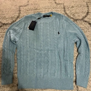 Ljusblå kabelstickad tröja Polo Ralph Lauren - Snygg ljusblå kabelstickad tröja från Polo Ralph Lauren med rund halsringning och klassisk broderad logga på bröstet. Tröjan har ribbade muddar vid ärmslut och nederkant. Perfekt för dig som gillar stilrena och tidlösa plagg. Helt ny aldrig använd. OBS! Lappen är borttagen (men fortsatt aldrig använd)