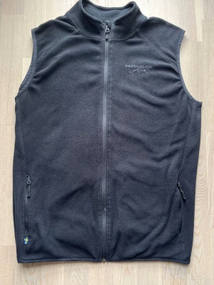 Swedemount väst - svart, XL - Geilo vest från Swedemount som är gjord i fleece med polyester som yttertyg. Västen har Swedemount-loggan på vänster bröst och dragkedja längs hela mitten. Plagget är i bra skick, men är använt och det finns små tecken på användning. Liten fläck vid dragkedjan och något nopprig vid halsen (se bilder).