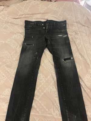 Svarta Dsquared2 jeans med slitningar - Säljer ett par svarta jeans från Dsquared2 med slitna detaljer och trasiga partier på benen. Jeansen har knappgylf, klassisk femficksmodell och en smal passform. Perfekta för dig som gillar en edgy och trendig stil.