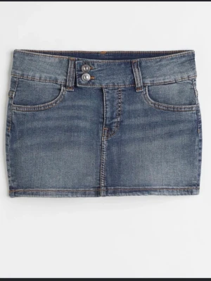 Blå lågmidjad jeanskjol med dubbla knappar - Säljer en klassisk blå lågmidjad jeanskjol med dubbla silverknappar i midjan och femficksmodell. Kjolen har en kort passform och är tillverkad i slitstarkt denimtyg. Perfekt att matcha med en croppad topp eller hoodie. Skulle säga att den sitter som en S!