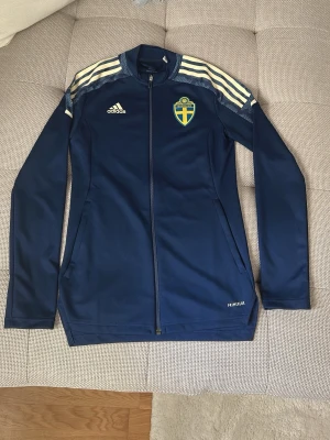 Blå Sverige träningsjacka Adidas - Mörkblå träningsjacka från Adidas med Sveriges landslagsemblem på bröstet och vita ränder på axlarna. Jackan har hel dragkedja, lång ärm och är tillverkad i funktionsmaterialet Primeblue. Perfekt för fotbollsträning eller sportiga dagar.