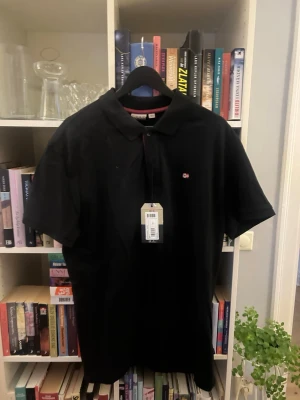Napapijri Pilétröja  - Klassisk svart pikétröja med krage och korta ärmar. På bröstet finns en liten broderad norsk flagga som detalj. Tröjan har en stilren look och är tillverkad i bomull för en bekväm känsla.