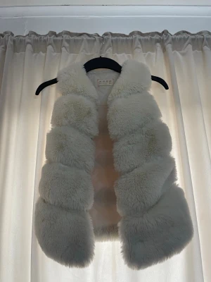 Vit fluffig pälsväst - Supersnygg vit pälsväst i fuskpäls med breda, horisontella paneler. Västen är ärmlös och har en mjuk, lyxig känsla som ger en trendig look. Perfekt att slänga över en hoodie eller tröja för extra stilpoäng. För barn 