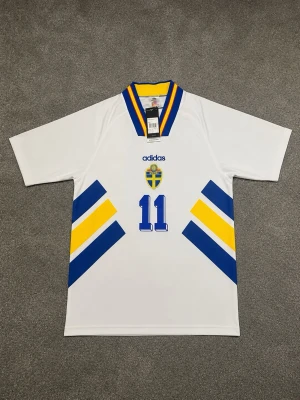 Svensk landslagströja Brolin #11 Adidas - Klassisk vit svensk landslagströja från Adidas med Brolin och nummer 11 på ryggen. Tröjan har blå och gula detaljer, V-ringad krage med blå och gula ränder samt Sveriges landslagsemblem på bröstet. Perfekt för dig som älskar fotboll och retrostil.