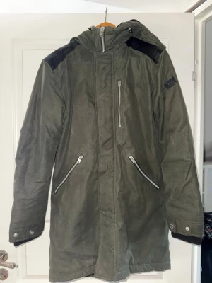 Olivgrön jacka med huva - Snygg olivgrön parkas med huva och svarta axeldetaljer. Jackan har tre dragkedjeförsedda fickor framtill och justerbara muddar med tryckknappar. Perfekt för kalla dagar och har en längre modell som skyddar mot vind och kyla.