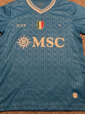 Napoli matchtröja ljusblå EA7 - Snygg ljusblå Napoli matchtröja från EA7 med vita detaljer och klubbmärke på bröstet. Tröjan har tryckta sponsorer som MSC och SORGESANA samt italienska mästarmärket. Tillverkad i lätt och ventilerande material, perfekt för fotboll eller träning.