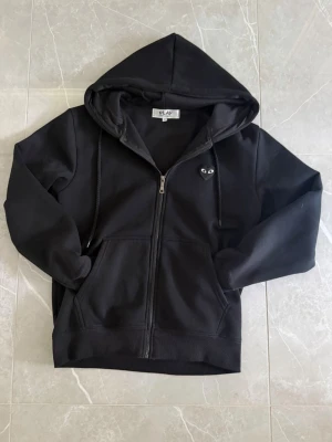 Svart hoodie med dragkedja och hjärtdetalj - Säljer en svart hoodie från PLAY i storlek M. Hoodien har dragkedja framtill, två fickor och en huva med dragsko. På bröstet sitter en liten svart hjärtdetalj med ögon. Tillverkad i mjukt bomullsmaterial, perfekt för en chill och stilren look.
