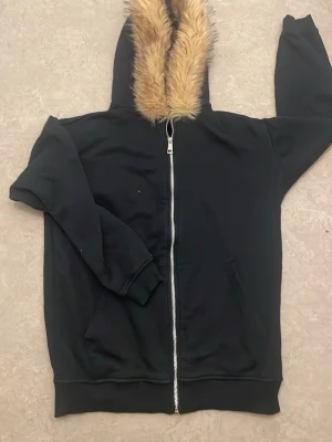 Svart hoodie med päls i huvan - Säljer en svart hoodie med dragkedja och stor huva fodrad med mjuk, ljusbrun fuskpäls. Hoodien har två fickor framtill och är gjord i ett mjukt, fleecefodrat material som känns riktigt skönt. Perfekt för dig som vill ha en chill och varm stil.