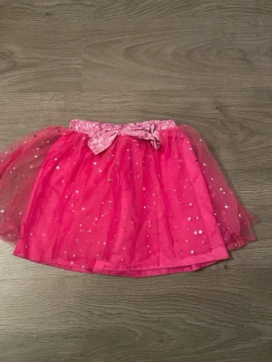 Rosa tyllkjol med rosett och glitter - Söt rosa kortkjol i tyll med glittriga stjärnor och en dekorativ rosett i midjan. Perfekt för dig som vill sticka ut lite extra med en lekfull och färgglad stil.