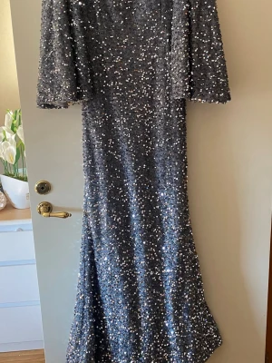 Glittrig långklänning med paljetter - Snygg långklänning i silver med glittriga paljetter över hela klänningen. Klänningen har v-ringning, vida ärmar och en elegant passform. Perfekt för dig som vill sticka ut på festen.
