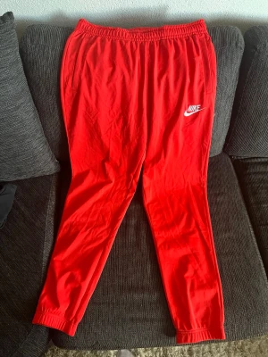 Röda mjukisbyxor från Nike - Snygga röda mjukisbyxor från Nike med vit logga på vänster ben. Byxorna har resår i midjan och vid bensluten samt två sidofickor. Perfekta för chill eller träning. Tillverkade i mjukt och bekvämt material.