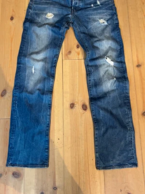 Blå slitna raka jeans - Säljer ett par blå jeans med raka ben och slitna detaljer på flera ställen. Jeansen har klassisk femficksdesign och är gjorda i denim. Perfekta för en avslappnad och trendig look.