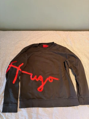 Hugo boss tröja - Svart sweatshirt med stor röd Hugo-logga i broderad stil som går över bröstet och ena ärmen. Tröjan har rund halsringning, långa ärmar och ribbade muddar vid ärmslut och nederkant. Perfekt för en trendig och avslappnad look.