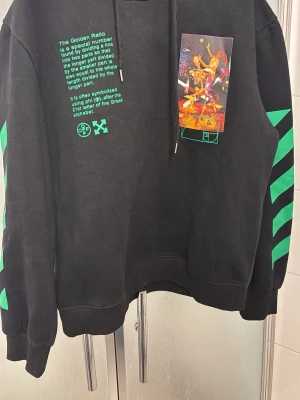 Off-White Golden Ratio hoodie svart/grön - Säljer en svart hoodie från Off-White med gröna detaljer och tryck. Framsidan har text och konstmotiv, medan ryggen har stort färgglatt print och texten 'Golden Ratio'. Ärmarna har gröna pilar. Klassisk huva med dragsko och ribbade muddar. Perfekt för dig som gillar streetwear.
