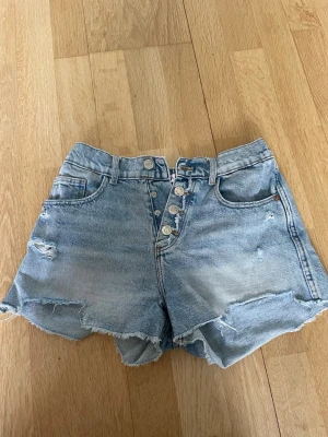 Ljusblåa högmidjade jeansshorts - Snygga ljusblå jeansshorts från Zara i storlek 164. Shortsen har knappar framtill och slitna detaljer.