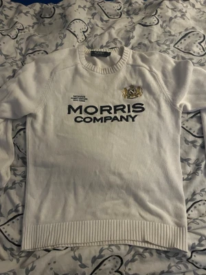 Vit stickad tröja Morris Company - Vit stickad tröja från Morris Company med svart text och ett broderat emblem på bröstet. Tröjan har rund halsringning, långa ärmar och ribbade muddar vid ärmslut och nederkant. Perfekt för dig som gillar klassisk och stilren design.
