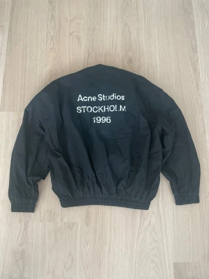 Svart vindjacka Acne Studios - Svart vindjacka från Acne Studios med tryck 'Acne Studios STOCKHOLM 1996' på ryggen. Jackan har dragkedja framtill, resår i midja och ärmslut samt hög krage. Tillverkad i lätt och tåligt material, perfekt för blåsiga dagar.