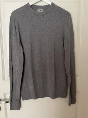 Grå rundhalsad tröja från H&M premium - En stilren grå tröja från H&M premium i slim fit-modell. Tröjan har rund halsringning, långa ärmar och ribbade muddar vid hals, ärmslut och nederkant. Perfekt basplagg för lager på lager och enkel att matcha med olika outfits.