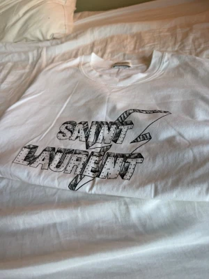 Saint Laurent t-shirt - Säljer en vit t-shirt från Saint Laurent med svart grafiskt tryck på bröstet. T-shirten har rund hals och korta ärmar, perfekt för en clean och stilren look. Materialet känns mjukt och skönt mot huden.