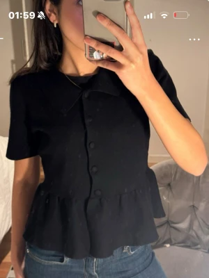 Svart blus med volang och krage - Snygg svart blus med korta ärmar, volang nertill och klassisk krage. Knäpps framtill med tygklädda knappar. Perfekt till jeans eller kjol för en trendig look. Materialet ser ut att vara mjukt och följsamt.