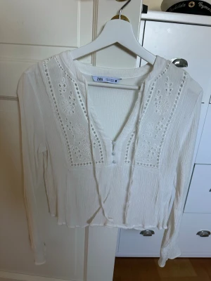 Vit blus med broderi från Zara - Superfin vit blus från Zara med broderade detaljer och knappar framtill. Blusen har långa ärmar, v-ringning och tunna knytband vid halsen. Materialet är lätt och luftigt, perfekt för en fräsch look. Broderierna ger en romantisk touch.