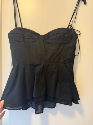Svart peplum från ZARA - Supersnygg svart linne-topp från ZARA med tunna axelband och peplumvolang. Toppen har markerade kupor, knytband och lätt transparent nederdel. Perfekt för dig som vill ha en trendig och feminin look.