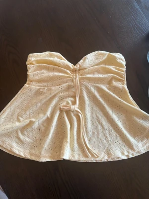 Gul axelbandslös tubtopp med snörning - Söt gul tubtopp i broderad bomull med dekorativ snörning framtill. Toppen är axelbandslös och har ett lätt utsvängd passform, perfekt för varma dagar. 