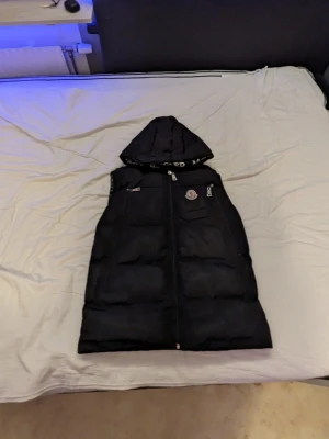 Svart dunväst med huva från Moncler - Snygg svart dunväst från Moncler med avtagbar huva och dragkedja framtill. Västen har Moncler-logga på bröstet, praktiska fickor med dragkedja och är perfekt för kyliga dagar. Klassisk pufferstil och exklusiv känsla.