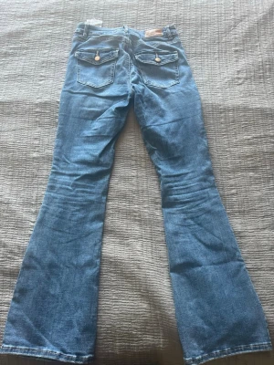 Blå bootcut jeans med fickdetaljer - Säljer ett par ljusblå bootcut jeans med två fickor med lock och knapp bak. Jeansen har klassisk femficksdesign och utsvängda ben. Perfekta för dig som gillar retrostil och vill ha en avslappnad look.