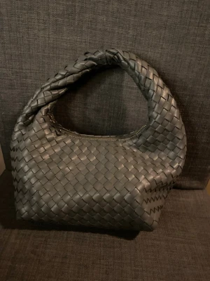 Svart flätad handväska från Gina Tricot - Trendig svart handväska från Gina Tricot med flätad design i konstläder. Väskan har en mjuk, rundad form och ett kort handtag som gör den enkel att bära. Stängs med dragkedja och har ett innerfack för småsaker. Är även sydd vid dragkedjan men det syns inte jätte mycket.