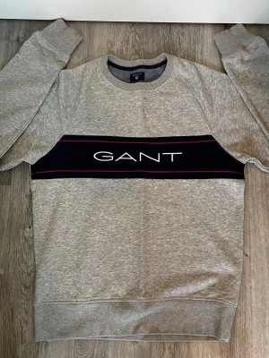 GANT sweater  - Snygg GANT tröja med en bred marinblå panel över bröstet och vit GANT-logga. Tröjan har rund hals, ribbade muddar och röda detaljer. Säljer pågrund av att den inte passar. Tröjan är i nyskick och den har varken defekter eller skador. Hör av er vid frågor, allt gott!