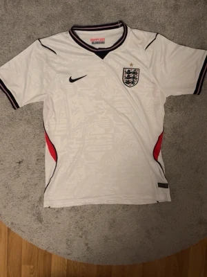 England Nike vit matchtröja - Säljer en vit England matchtröja från Nike med svart logga och landslagets emblem på bröstet. Tröjan har svarta och röda detaljer vid ärmslut och sidor samt diskret mönster i tyget. Perfekt för fotbollsfans som vill representera sitt lag.
