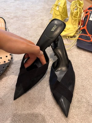 Svarta spetsiga pumps från Zara - Säljer ett par svarta pumps från Zara med spetsig tå och låg klack. Skorna har en snygg korsad design framtill i textilmaterial och öppen häl. Perfekta för dig som gillar stilrena och trendiga skor.