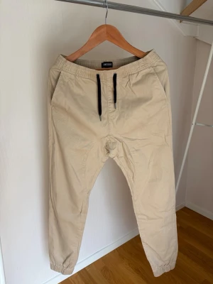 Beiga chinos med resår och snörning, joggers. - Snygga beige mjukisbyxor från Jack & Jones med elastisk midja och svarta dragsnören. Byxorna har två sidofickor fram och två bakfickor, varav en med knapp. Avsmalnande ben med resår vid benslut. Perfekta för en avslappnad stil.