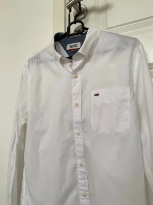 Vit skjorta från Tommy Hilfiger - Klassisk vit skjorta från Tommy Hilfiger med knappstängning framtill och en bröstficka med den ikoniska loggan. Skjortan har lång ärm och normal passform, perfekt till många olika stilar. 