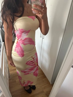 Beige långklänning med rosa blommor - Trendig axelbandslös långklänning i beige med stora rosa blommor. Klänningen har en tight passform och är perfekt för dig som vill sticka ut. Materialet känns mjukt och följsamt mot kroppen.