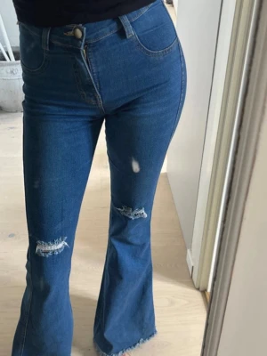 Blå utsvängda jeans med slitningar - Säljer ett par blå jeans med hög midja och utsvängda ben. Jeansen har slitna detaljer vid knäna och råa kanter nertill. Klassisk femficksmodell i stretchigt denimtyg. Perfekta för en trendig och avslappnad look.