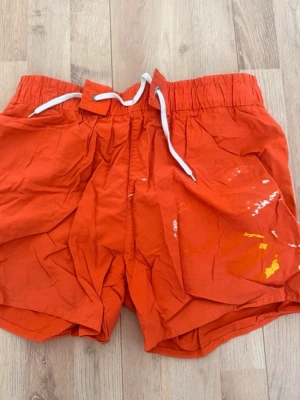 Orange loose shorts med snörning - Orange shorts med vit snörning i midjan och elastisk resår. Shortsen har en loose passform och är tillverkade i ett lätt syntetmaterial. Unik touch med färgstänk i vitt och gult på ena benet, perfekt för sommaren.