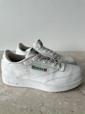 Vita Reebok Club C 85 sneakers storlek 37 - Klassiska vita Reebok Club C 85 sneakers i läder med gröna detaljer och logga på sidan och hälen. Skorna har lågt skaft, perforerad tå och vit sula. Perfekta för en clean och stilren look.