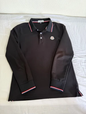 Svart Moncler pikétröja långärmad - Snygg svart långärmad pikétröja från Moncler med klassisk krage och knappar framtill. Tröjan har Moncler-logga på bröstet och detaljer i rött, vitt och blått på krage och ärmslut. Perfekt för en stilren och sportig look.