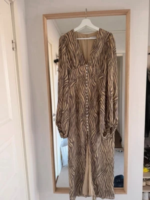 Adoor klänning zebra  - Säljer en långklänning med zebramönster i beige och brunt. Klänningen har v-ringning, långa puffärmar och knappar hela vägen framtill. Perfekt för dig som vill sticka ut med ett djurmönster och gillar en avslappnad men trendig look.