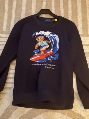 Svart sweatshirt Polo Bear Ralph Lauren - Cool svart sweatshirt från Polo Ralph Lauren med ikoniska Polo Bear som surfar på framsidan. Tröjan har rund hals, långa ärmar och tryckt motiv i färg. Perfekt för dig som gillar streetstyle och vill sticka ut med en unik print.