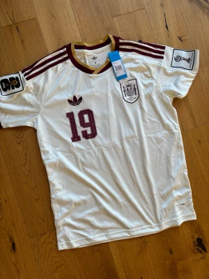 Vit Spanien matchtröja Adidas - Säljer en vit Spanien matchtröja från Adidas med vinröda detaljer och nummer 19. Tröjan har Lamine Yamal tryckt på ryggen, officiella klubbmärken och snygga ränder på axlarna. Tillverkad i lätt och ventilerande material, perfekt för fotboll eller träning. Vm tröja helt oanvänd 